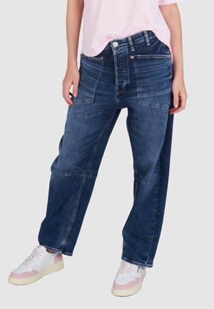 Donkerblauwe denim jeans met een losse fit, zichtbare stiksels en grote voorzakken. Gecombineerd met witte sneakers met roze accenten.