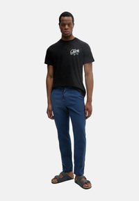 Camiseta de algodón negra con logo blanco "Guess", pantalones azules y sandalias marrones. El conjunto presenta un ajuste relajado y un diseño casual.