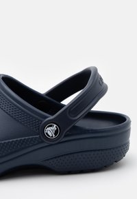 Temno modri klobuki z teksturirano površino, s slingback trakom in okroglim logotipom. Narejeni iz materiala, ki spominja na gumo, z perforiranim podplatom.