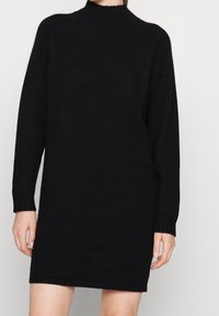 Robe tricotée noire avec un col montant, des manches longues et des poignets côtelés. Texture douce, design à mi-cuisse, style minimaliste.