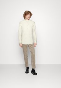 Camicia beige chiaro a maniche lunghe con colletto alla mandarina e tre bottoni, abbinata a pantaloni in corduroy beige e scarpe nere con lacci.