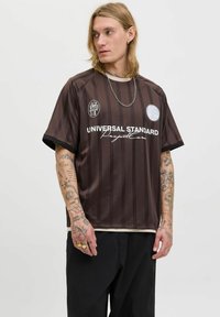 Ung mand med tatoveringer bærer en brunstribet "Universal Standard Project Core" skjorte og sorte bukser, mens han ser til venstre mod en ensfarvet baggrund.