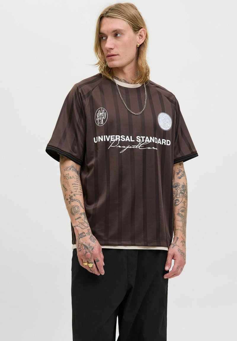 Ung mand med tatoveringer bærer en brunstribet "Universal Standard Project Core" skjorte og sorte bukser, mens han ser til venstre mod en ensfarvet baggrund.