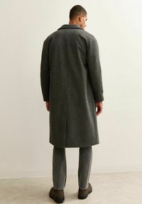 Cappotto di lana grigio con colletto scollato, apertura posteriore singola e maniche con tre bottoni. Indossato sopra pantaloni grigi e stivali marroni.