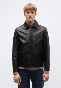 Fekete bőrdzseki hegyes gallérral, elülső cipzárral és oldalzsebekkel. Sima textúrájú és testre szabott fazonú, bézs ing fölött viselve.