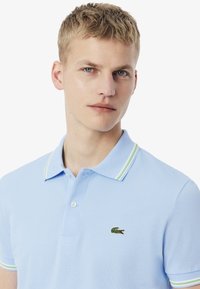 Ljusblå piké skjorta i texturerad tyg, med en grön krokodil-logotyp och treknapps knäppning med kontrasterande randig krage.