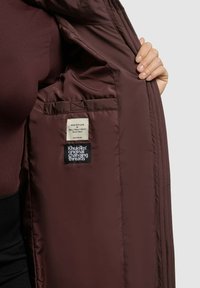 Unutrašnjost burgundske jakne sa izolacijom s platnenom etiketom koja glasi "NAPRAVLJENO S LJUBAVLJU" i oznakama brenda. Vidljiva glatka tekstura i šivani detalji.