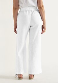 Witte linnen broek met wijde pijpen en een elastische tailleband, met een gestructureerde stof en een losse fit. Schoenen zichtbaar bij de zoom.