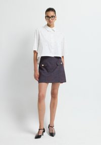 Witte cropped blouse met gouden accenten, gecombineerd met een marineblauwe minirok met patroon, voorzien van zakken aan de voorkant en gouden knopen. Zwarte T-strap hakken.