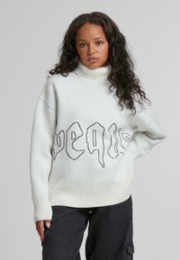 Weißer, oversized Turtleneck-Pullover mit schwarzem Grafiktext. Rippbündchen und Saum. Weiches Material und entspannte Passform. Getragen mit dunklen Hosen.