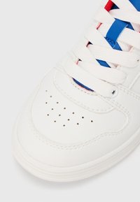 Pantof sport alb din piele netedă. Prezintă șireturi textile în alb, roșu și albastru, cu perforații pe căptușeala degetelor și cusături contrastante.