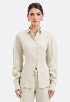 Kvinde med mørkt hår iført beige skjorte med lange ærmer og matchende bukser, stående mod en ensfarvet hvid baggrund.