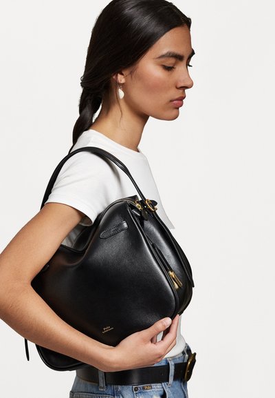 Juodas odinis rankinis krepšys su kreivinės dizainu, su auksiniais aksesuarais ir užtrauktuku. Laikomas rankoje, turi lygų tekstūrą, turi subtilų logotipą.