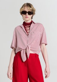 Persona che indossa occhiali da sole tartarugati, una collana a strati con perline rosse, pantaloni rossi e una maglietta a maniche corte a righe bianche e rosse annodata.