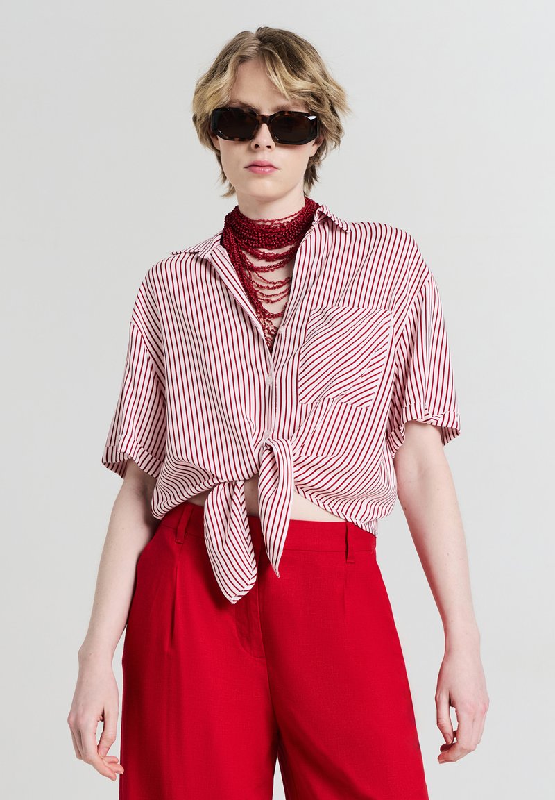 Persona che indossa occhiali da sole tartarugati, una collana a strati con perline rosse, pantaloni rossi e una maglietta a maniche corte a righe bianche e rosse annodata.