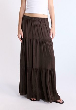 Ženska, oblečena v beli kratki top in dolgo, večplastno temno rjavo krilo z naborki, stoji z rokami sproščeno ob telesu.