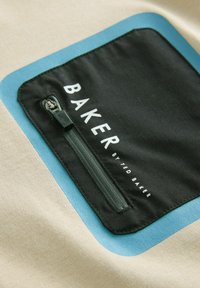 Beige tyg med en svart ficka med dragkedja, markerad med en blå kant. Texten lyder "BAKER av TED BAKER" i vitt.