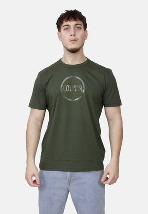 Giovane uomo in piedi di fronte, indossa una maglietta verde con il logo COLMAR rotondo e jeans azzurri chiari su uno sfondo semplice.