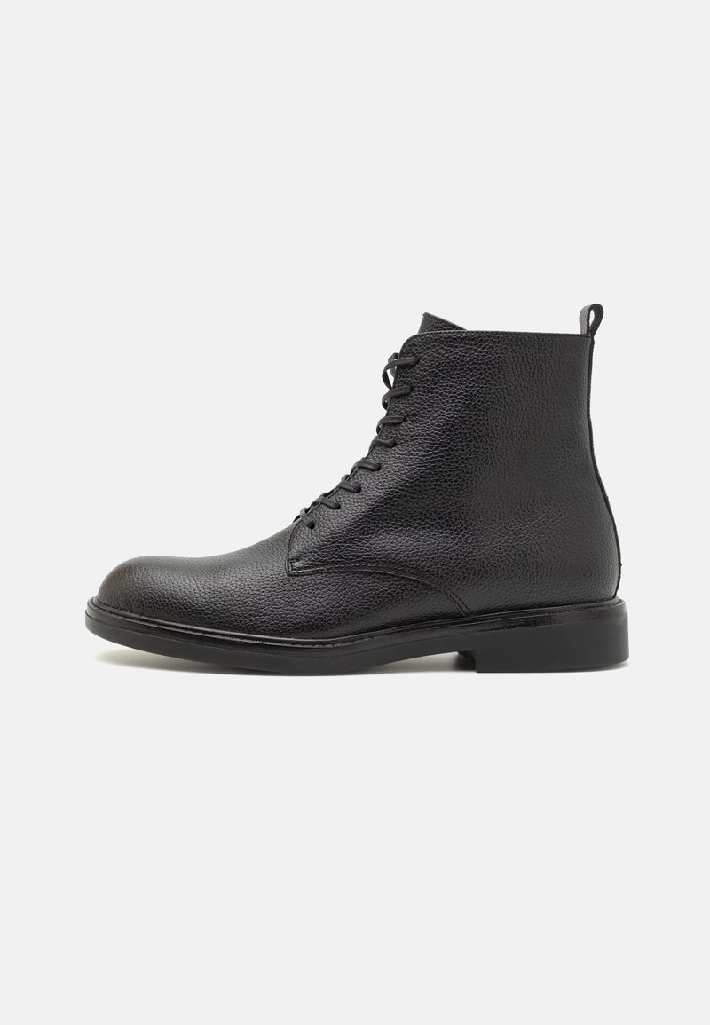 Bottine en cuir noir à surface texturée, fermeture à lacets sur le devant, languette au talon et semelle basse empilée sur fond blanc.