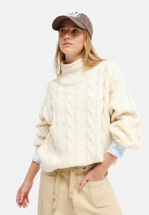 Frau mit beiger Kappe, cremefarbenem Zopfmuster-Rollkragenpullover und beiger Hose, eine Hand in der Tasche vor einem einfarbigen Hintergrund.