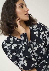 Blusa nera a fantasia floreale con fiori bianchi; presenta un collo a V, maniche puff e polsini arricciati realizzati in un tessuto leggero.
