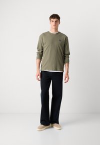 Olijfgroene geribbelde lange mouw shirt met witte binnenkraag, gecombineerd met donker wijdvallende jeans en lichtgekleurde sneakers.
