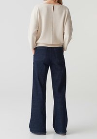 Pull crema a coste con dettaglio a chiave sulla schiena, abbinato a jeans svasati blu scuro con tasche posteriori e una finitura liscia.