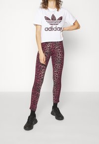 Woman standing wearing white Adidas crop top with leopard logo, pink leopard print leggings, black socks, and black sneakers.