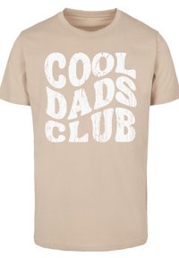 Camiseta beige de algodón con mangas cortas. Presenta un gran gráfico en blanco con la frase "CLUB DE PAPÁS GENIALES" en una fuente gruesa y desgastada.