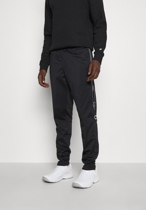 Mężczyzna w czarnych dresowych spodniach Champion z logo na bokach, czarnej koszulce z długim rękawem i białych sneakersach sportowych stoi na tle jednolitym.