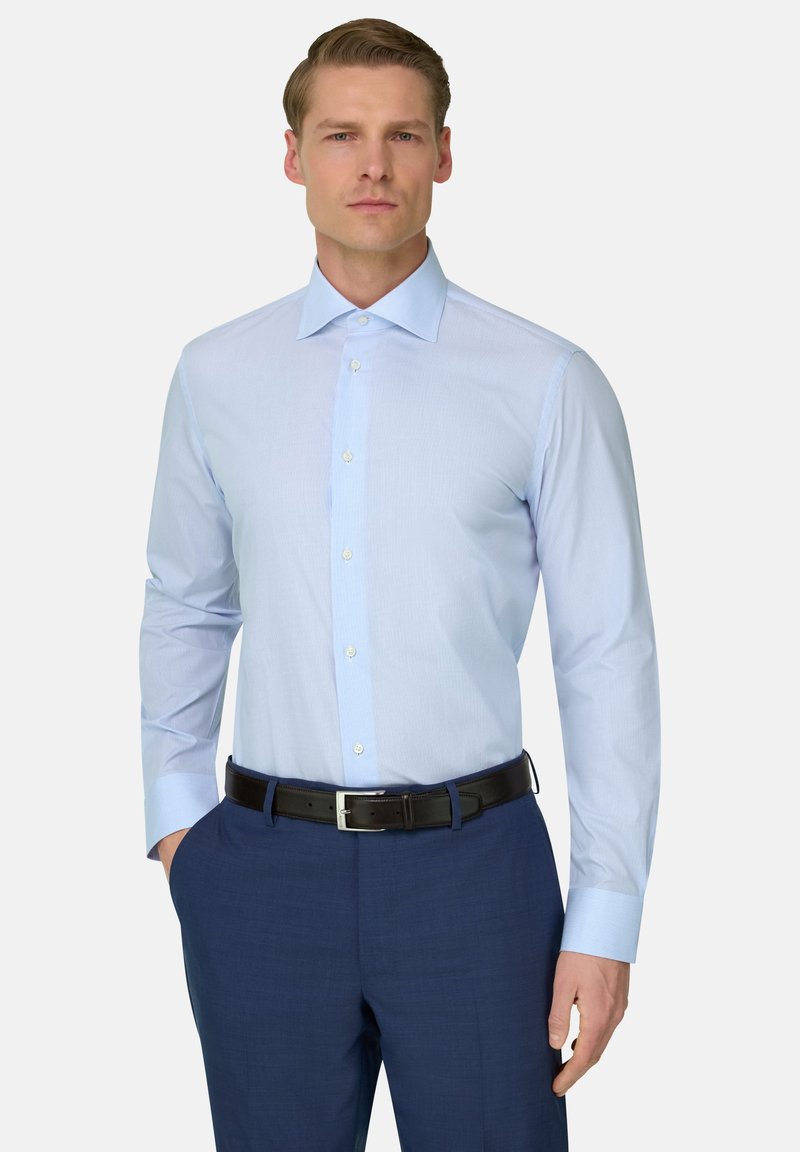 Homme portant une chemise habillée bleu clair rentrée dans un pantalon bleu marine avec une ceinture noire, debout une main dans la poche devant un fond uni.