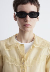 Yellow sheer button-up shirt with lace details across the chest, paired with a white crew neck shirt underneath and black rectangular sunglasses.