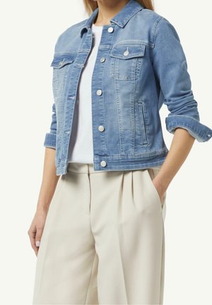 Femme portant une veste en jean bleu clair sur une chemise blanche avec un pantalon beige taille haute, une main dans la poche, manches retroussées.