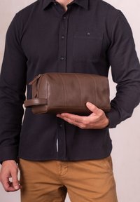 Man wearing black shirt and brown pants holds a brown leather zippered toiletry bag with side strap at waist level.