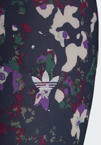 Mörk tyg med abstrakt blommönster i lila, grönt och krämfärgat, med en vit broderad Adidas trefoil-logotyp.
