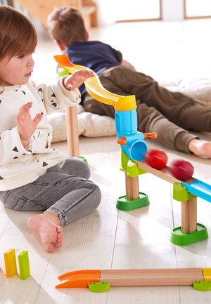 Bunter Spielzeug-Murmelbahn mit einer gelben Rutsche, blauem Trichter, grünen Stützen und roten Kugeln, auf einem hellen Holzboden aufgebaut.