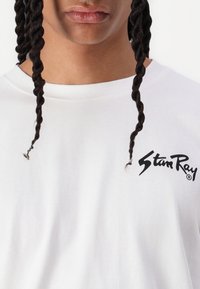 Close-up of a person with two braided strands of hair wearing a white shirt with "Stan Ray" logo on the chest.