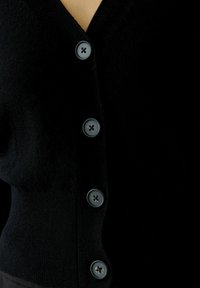 Schwarze Strickjacke aus weichem Stoff, mit V-Ausschnitt, sechs grauen Vorderknöpfen im Vierloch-Muster und einemtaillierten Bund.