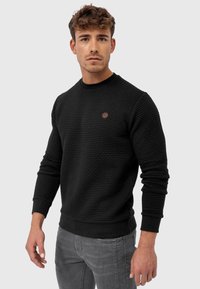 Schwarzer Pullover mit Struktur, rundem Ausschnitt, gerippten Bündchen und Saum, mit kleinem Logo auf der Brust. Kombiniert mit grauen Jeans.
