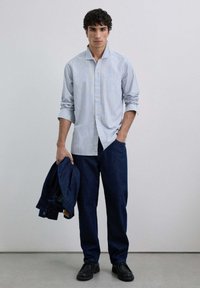 Camisa de cuadros en azul claro y blanco, combinada con pantalones de mezclilla azul oscuro. El modelo sostiene una chaqueta de mezclilla, lleva zapatos negros y un fondo neutro.