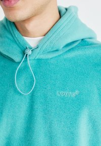 Turkio spalvos flisinė bliuzono su raišteliu uždarymo, minkšta tekstūra ir siuvinėtas Levi's logotipas atitinkama spalva. Kapšas turi aukštą apykaklę.