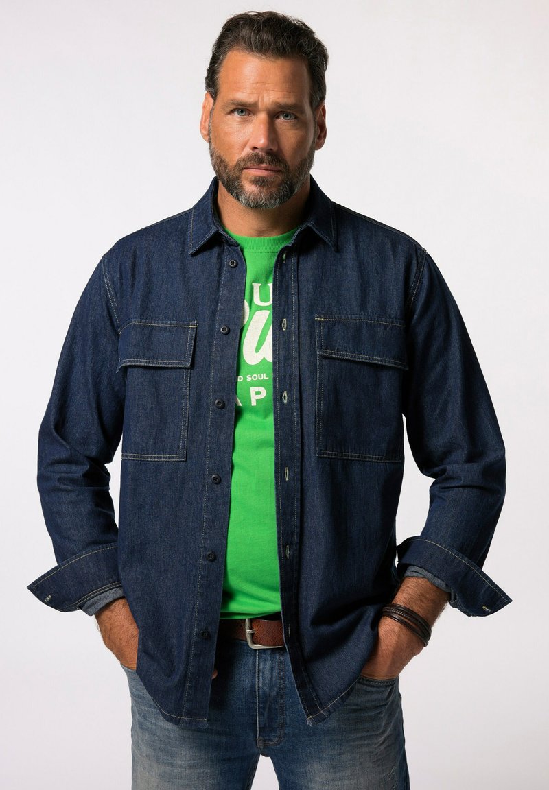 Camicia di jeans con un lavaggio blu scuro, due tasche frontali, bottoni sul davanti, indossata sopra una t-shirt grafica verde, abbinata a jeans chiari.