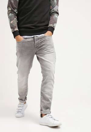 Jeans en denim gris clair, coupe slim avec un léger délavage, associés à des baskets blanches. Le modèle porte un sweatshirt noir avec des manches à motifs.