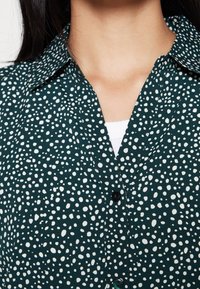 Camicia verde con un motivo a pois bianchi, dotata di colletto e bottoni neri, realizzata in un morbido tessuto strutturato.