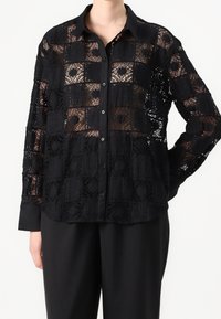 Svart spetsblus med rutmönster, knappstängning, långa ärmar och svart krage samt manschetter i helsvart. Halvgenomskinlig design.