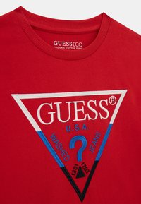 Punainen orgaaninen puuvillapaita, jossa on valkoinen ja sininen kolmion muotoinen logo, jossa lukee "GUESS®", "WASHED", "JEANS" ja koristeellinen kysymysmerkki-motiivi.