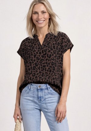 Femme souriante portant un chemisier à manches courtes noir et marron à imprimé léopard, rentré dans un jean bleu clair, tenant un sac tissé dans une main.