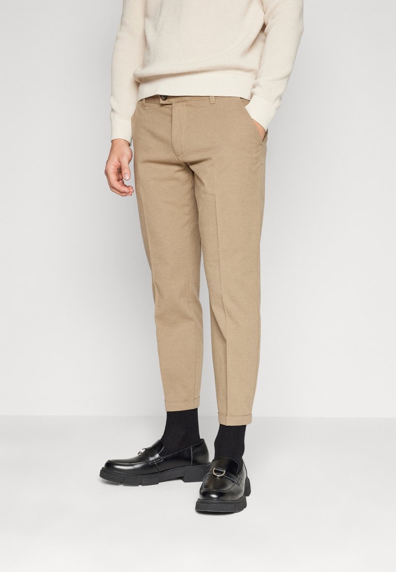Beige chinos with a tapered fit, featuring a smooth texture, front pockets, and rolled cuffs, paired with black shoes and dark socks.