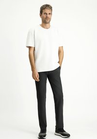 T-shirt bianco a maniche corte, colletto a giro, abbinato a pantaloni neri slim-fit. Il modello indossa scarpe sportive su uno sfondo neutro.