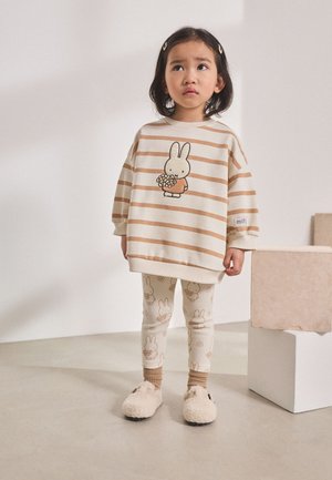 Unge barn i beige og brune stribede oversized sweatshirt med kanin, der holder blomster, matchende leggings og bløde hjemmesko, stående indendørs.